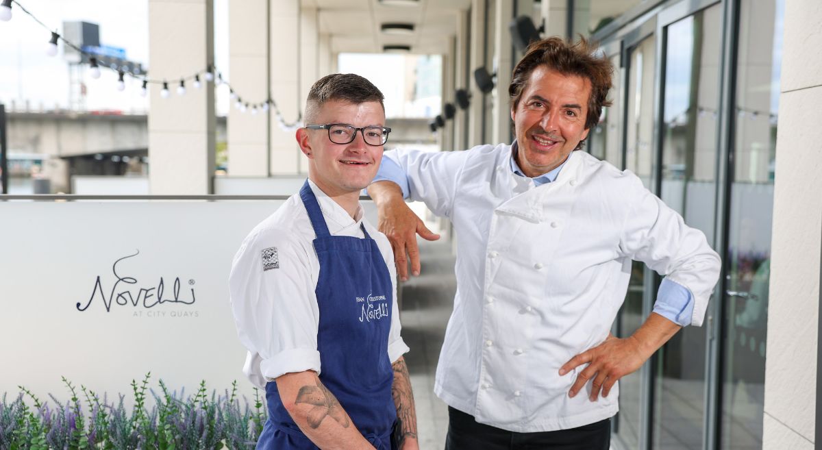 Two male chefs wearing chef whites, one with a blue apron, pictured outside Novelli's Restaurant